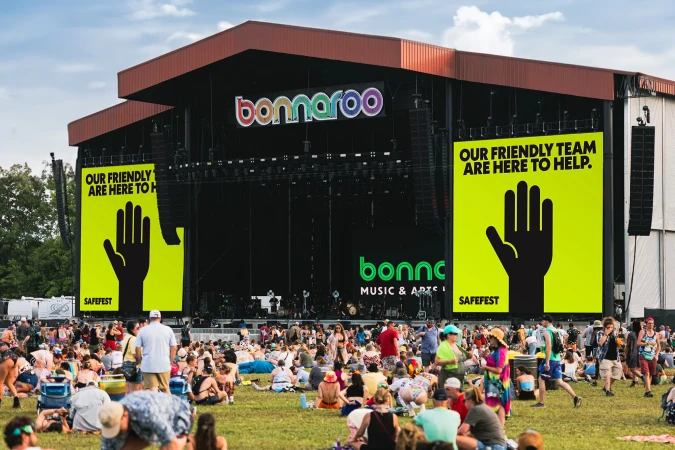 Crowd of people gathered at the Bonnaroo music festival with a large stage and signage in the background on a sunny day.