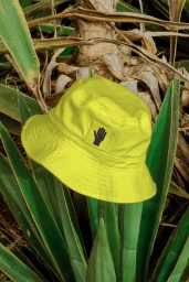 Bright yellow bucket hat with a black hand logo patch suspended amidst large pointed green agave leaves.