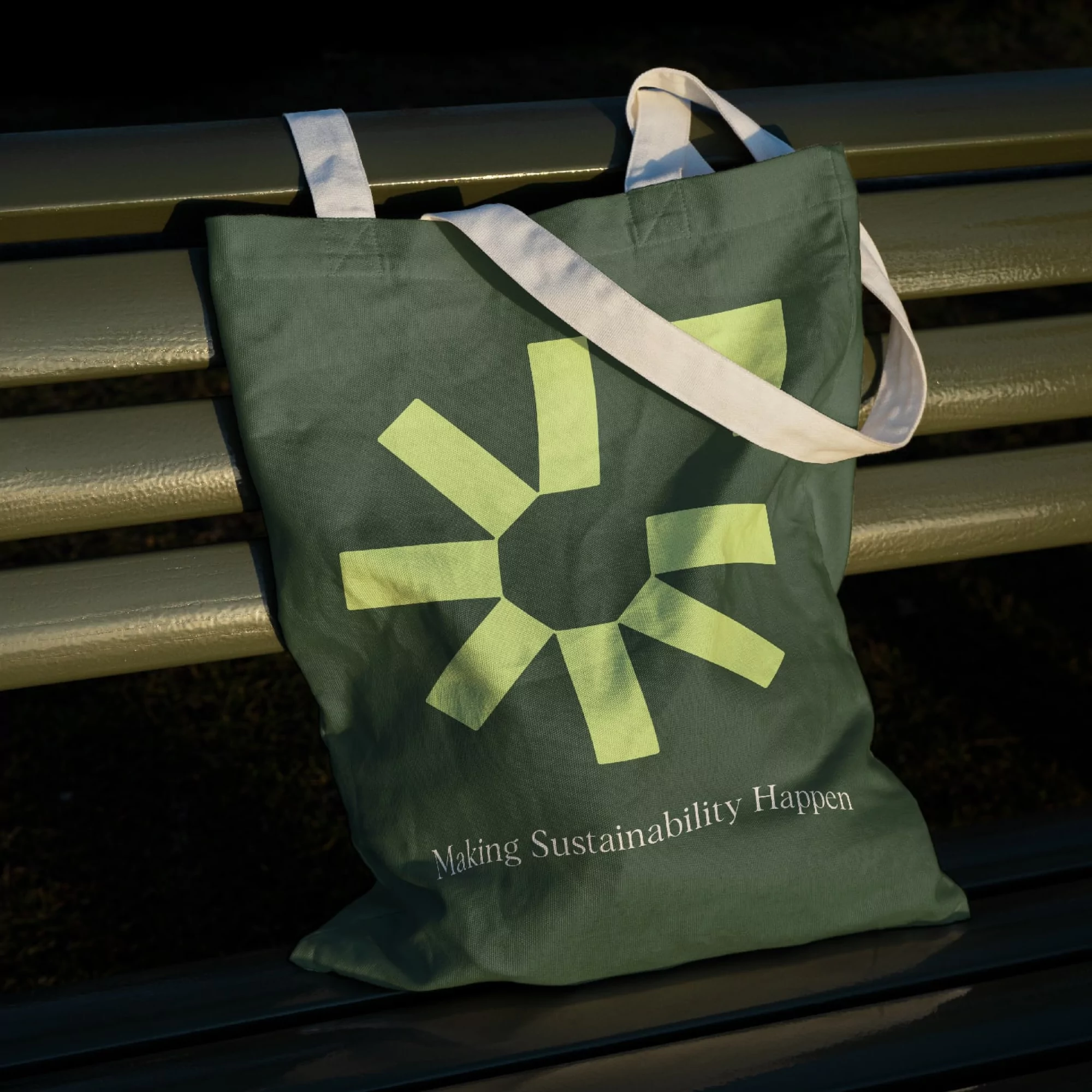 Green reusable tote bag with a graphic sun and text "Making Sustainability Happen" resting on a slatted green bench outdoors.