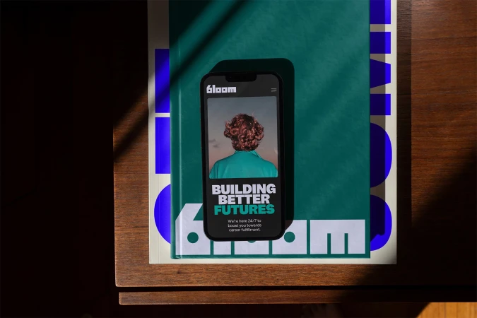 Mobile phone displays 'Bloom' website on a book, promoting career fulfillment, with a warm color palette and shallow depth of field.