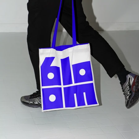 A person walking carries a white tote bag with blue graphic design, wearing dark pants and silver running shoes, indoors against a white wall.