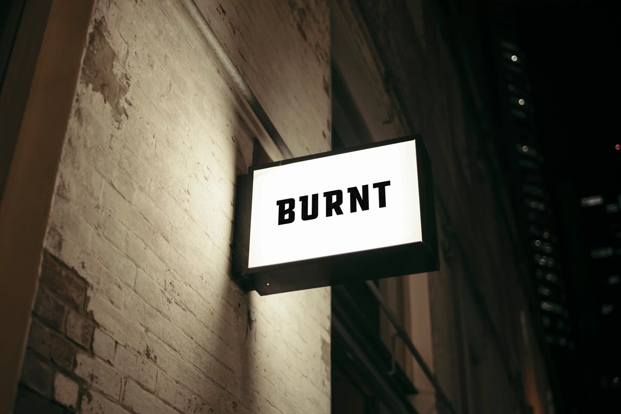 Illuminated sign displays the word 'BURNT' in bold letters against a textured brick wall at night, with a blurred cityscape in the background.