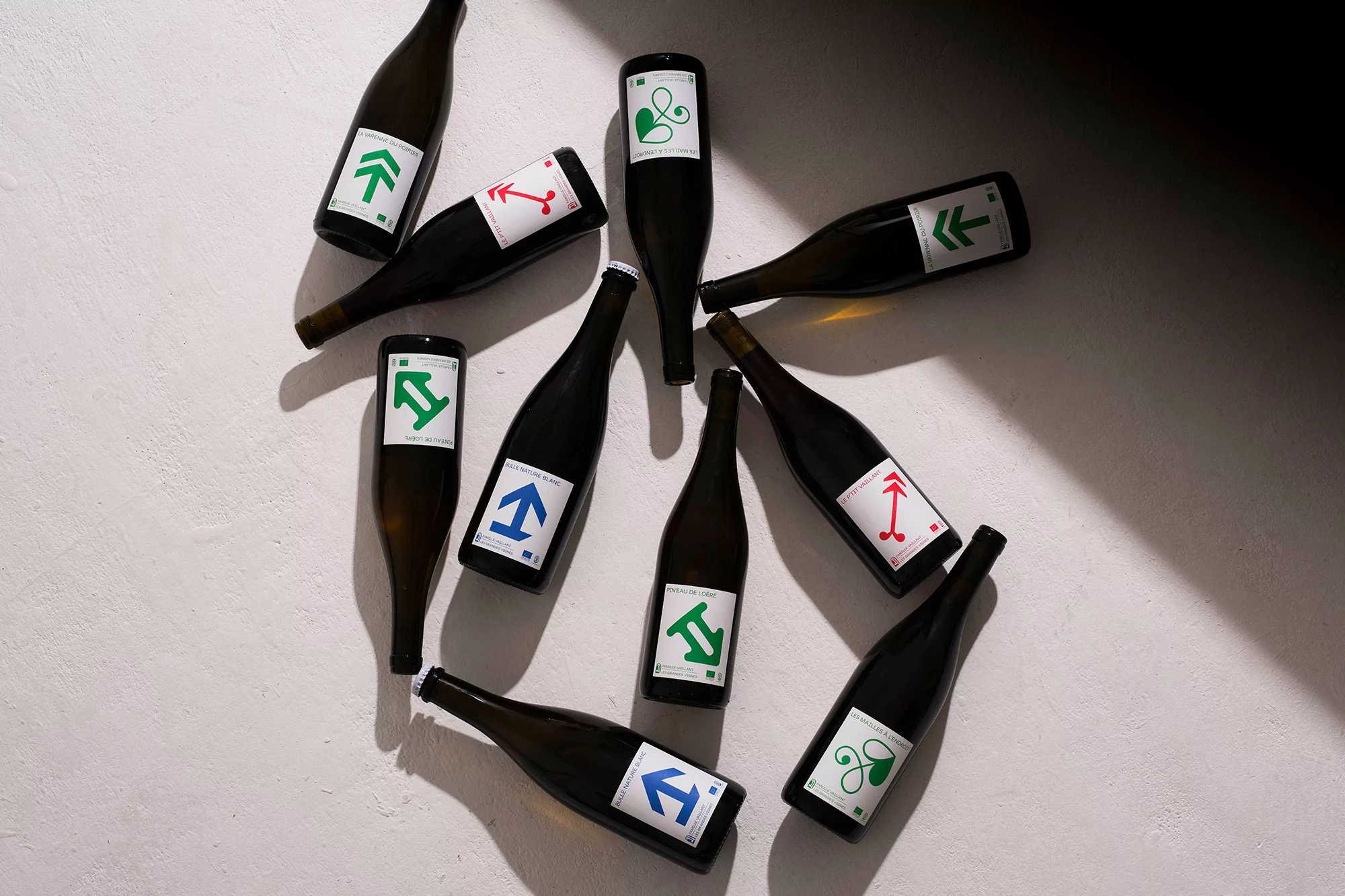 Arrangement of wine bottles with colorful, geometric label designs on a textured white surface, with natural light and soft shadows.