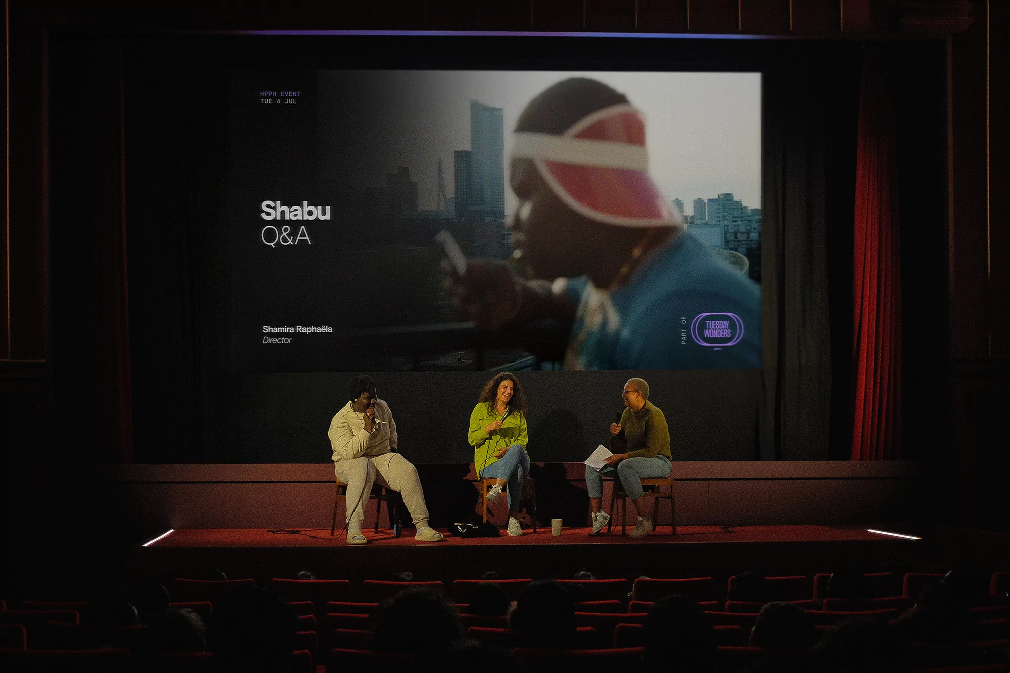 Three individuals on stage during a Q&A session in a darkened theatre, with a video of Shabu playing in the background.