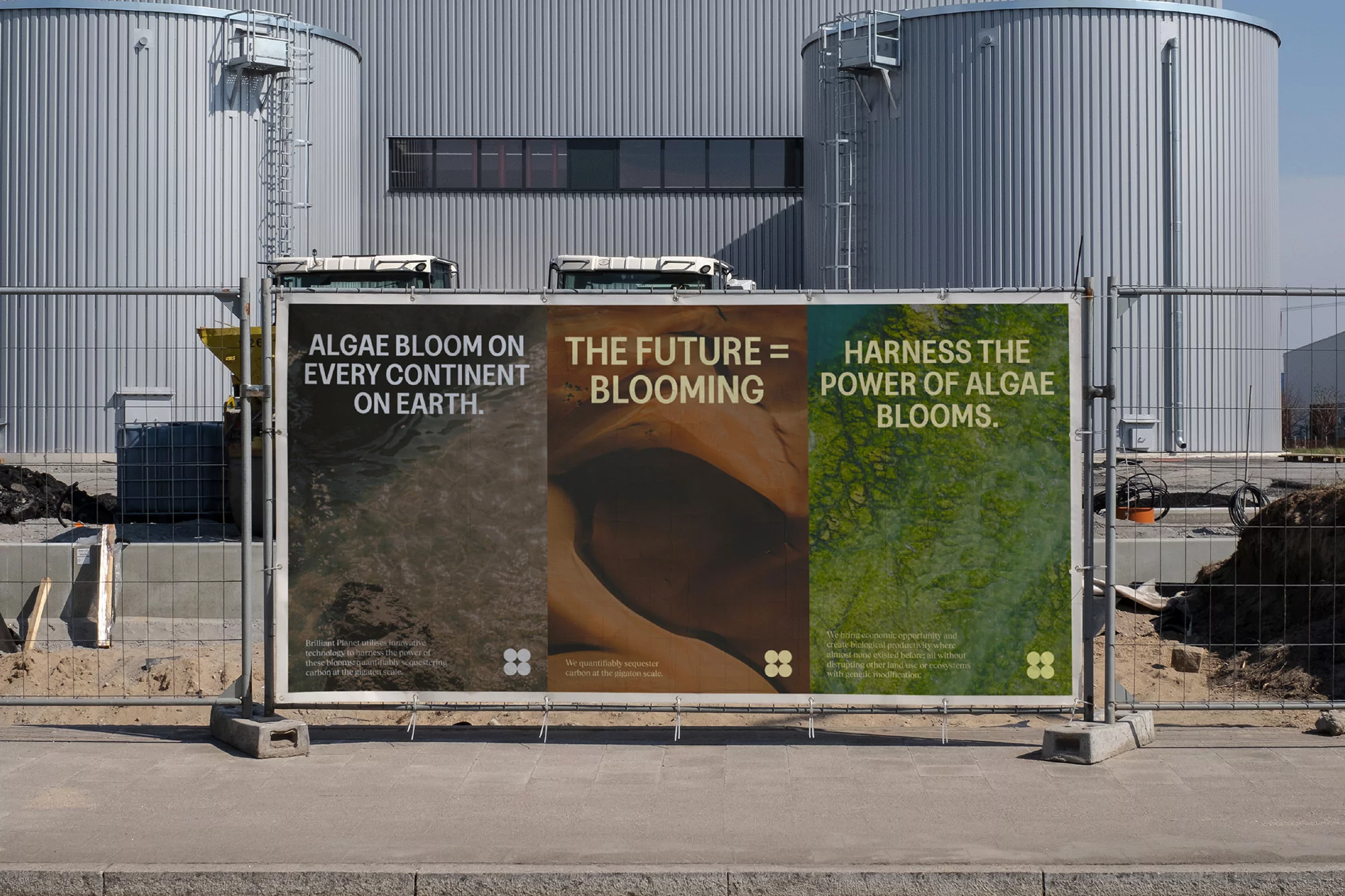 Three advertising posters on a metal fence display messages about algae blooms against a backdrop of large industrial tanks.