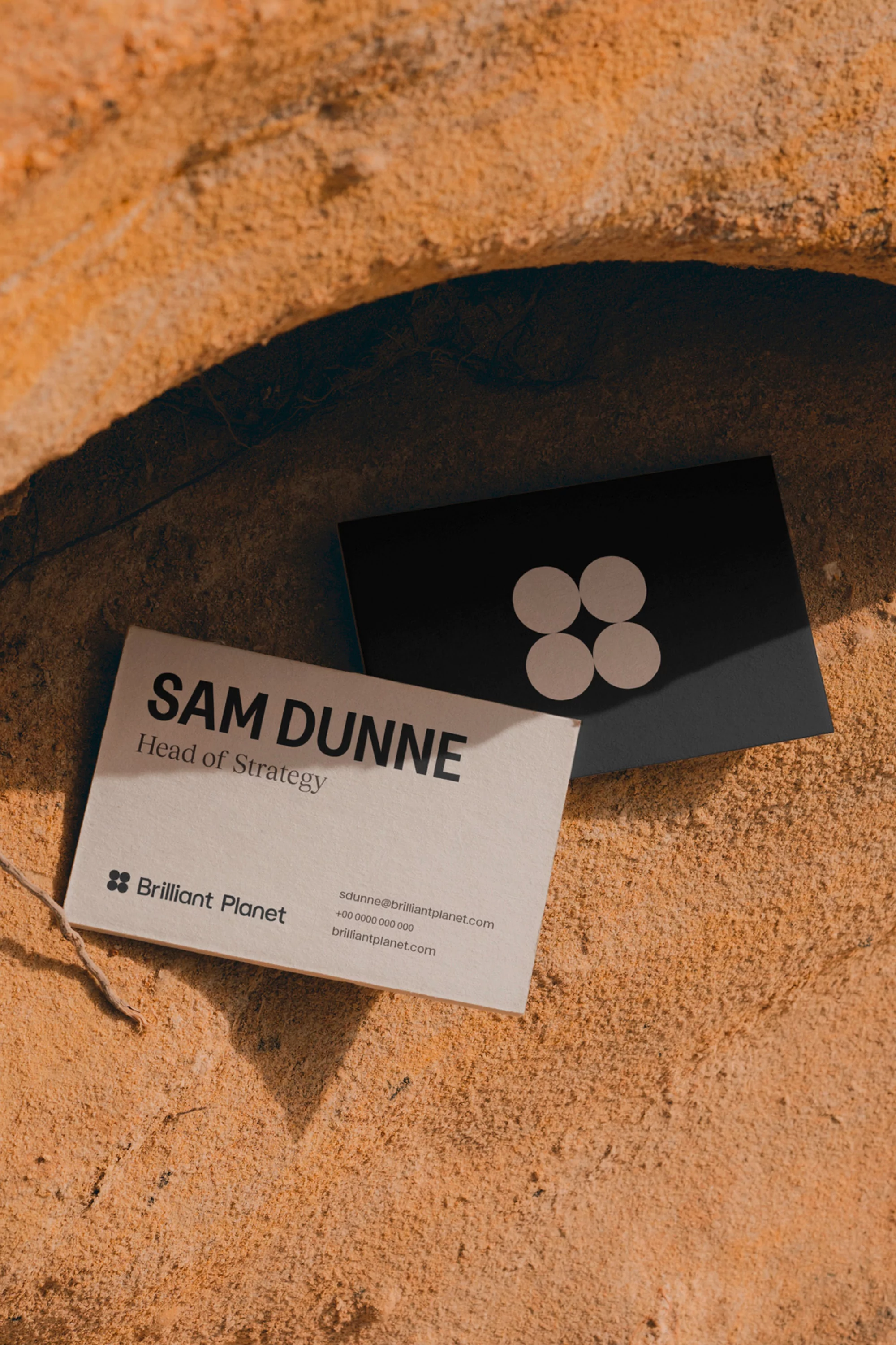 Two business cards, one white with black text, the other black with white circles, sit on a textured tan surface in natural light.