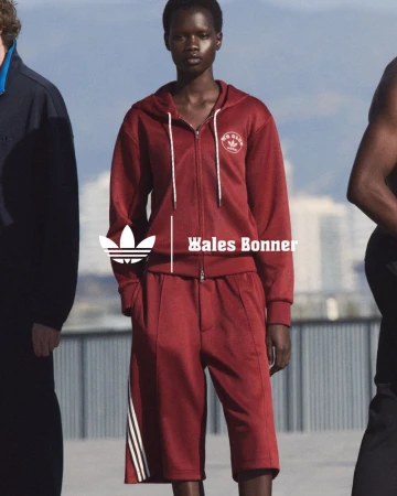 Two adult models showcase Adidas x Wales Bonner maroon tracksuit with stripes, set against a blurred urban backdrop with natural lighting.