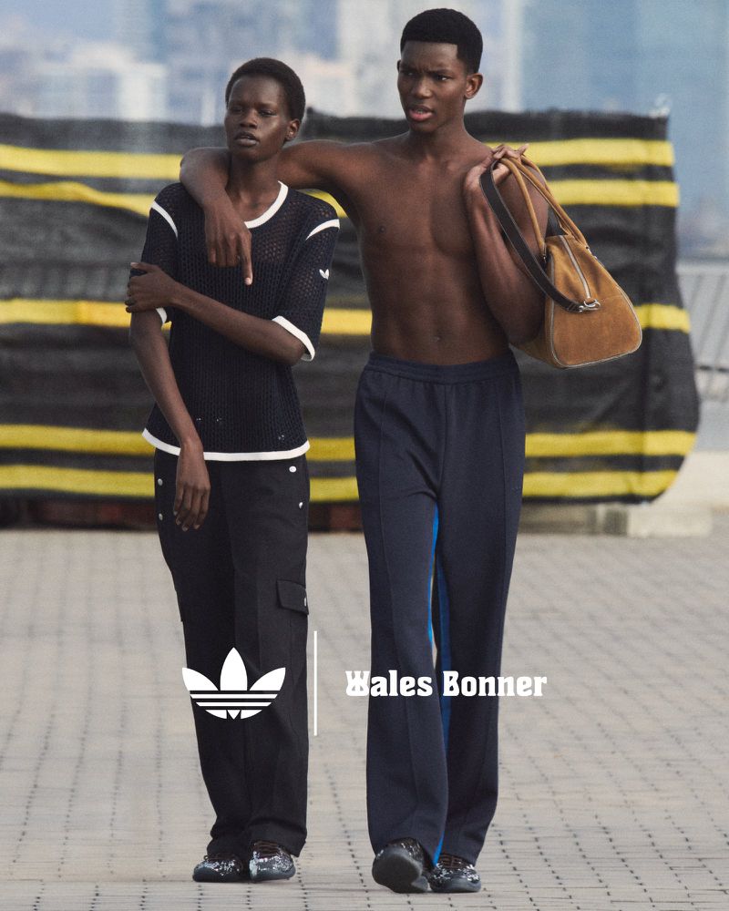 Two Black fashion models in Wales Bonner and Adidas clothing walk on a city street, one shirtless with bag, in medium outdoor shot.