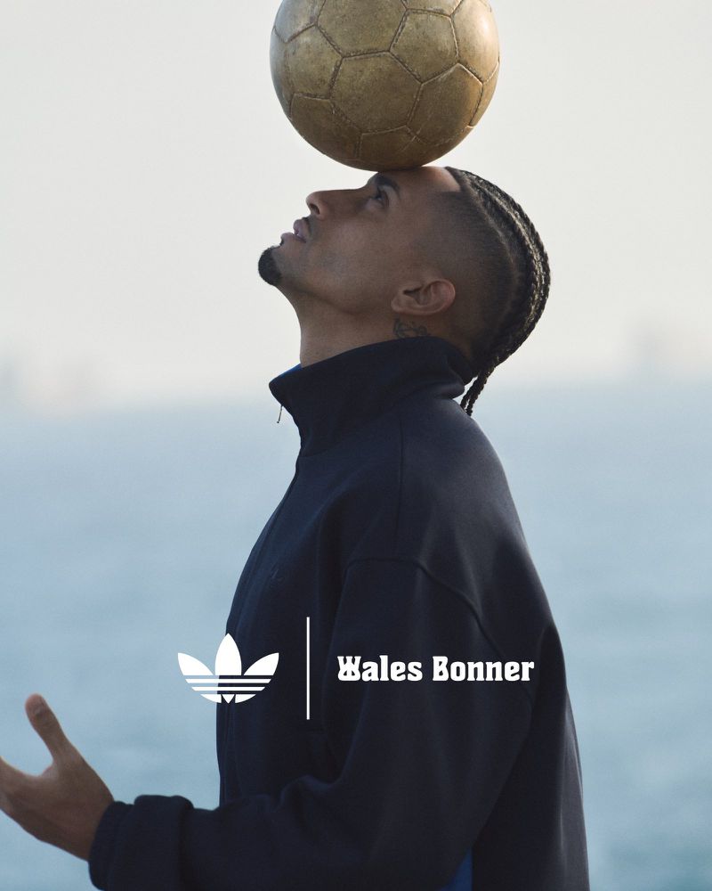 Portrait of a man balancing a soccer ball on his head outdoors. He is wearing an Adidas track jacket. Water visible in the background.