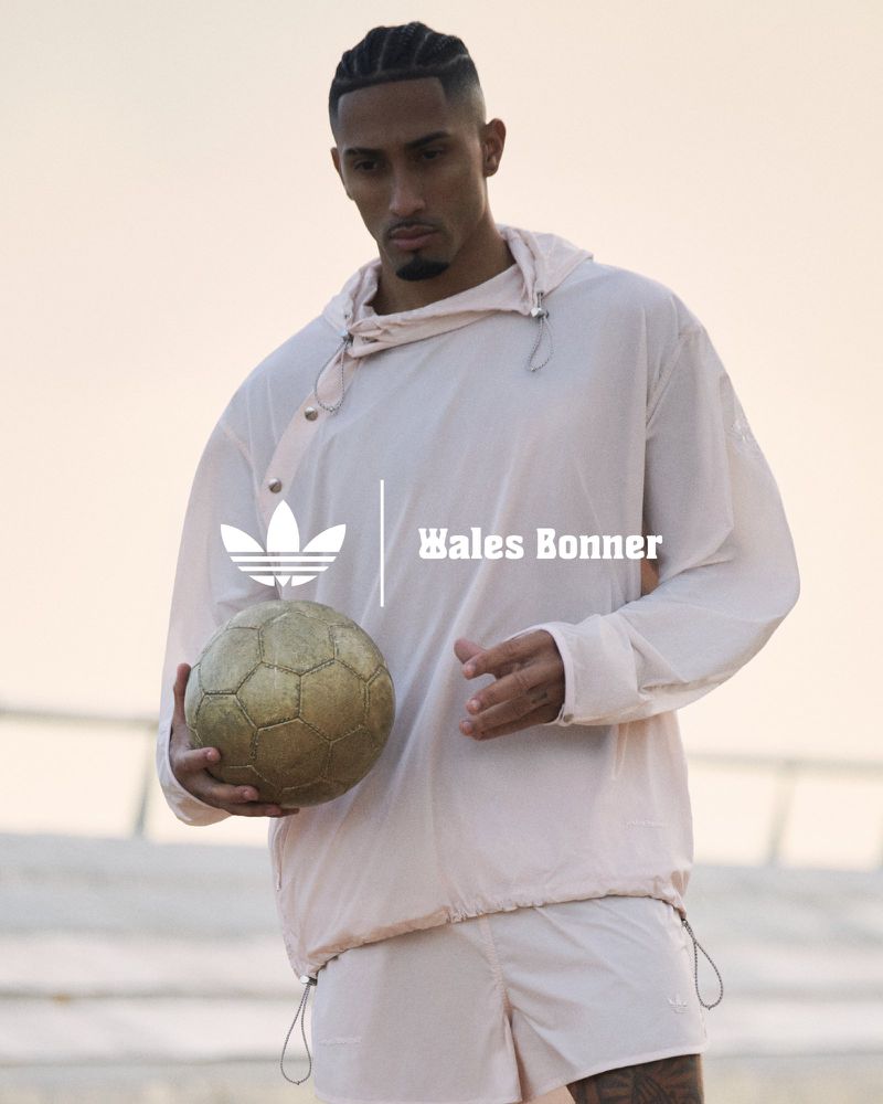 Athletic Black man in Adidas x Wales Bonner campaign, wearing hooded jacket and shorts, holding gold soccer ball outdoors in golden light.