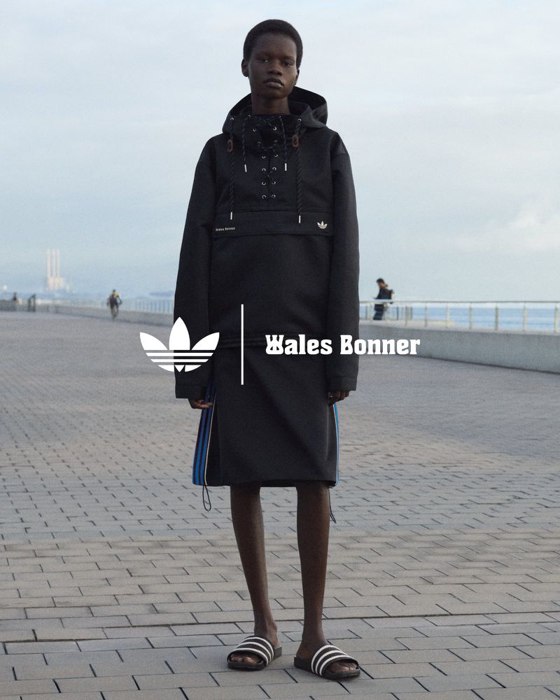 Fashion shot of a woman in a black hooded jacket and skirt, standing on a paved walkway, with a city skyline in the background.