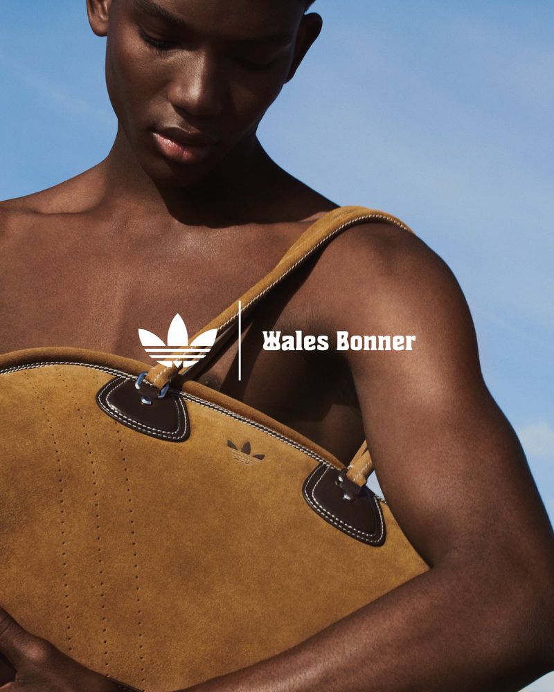 Adult male carrying a tan suede duffel bag by Adidas x Wales Bonner, outdoors against a light blue sky in a fashion shot.