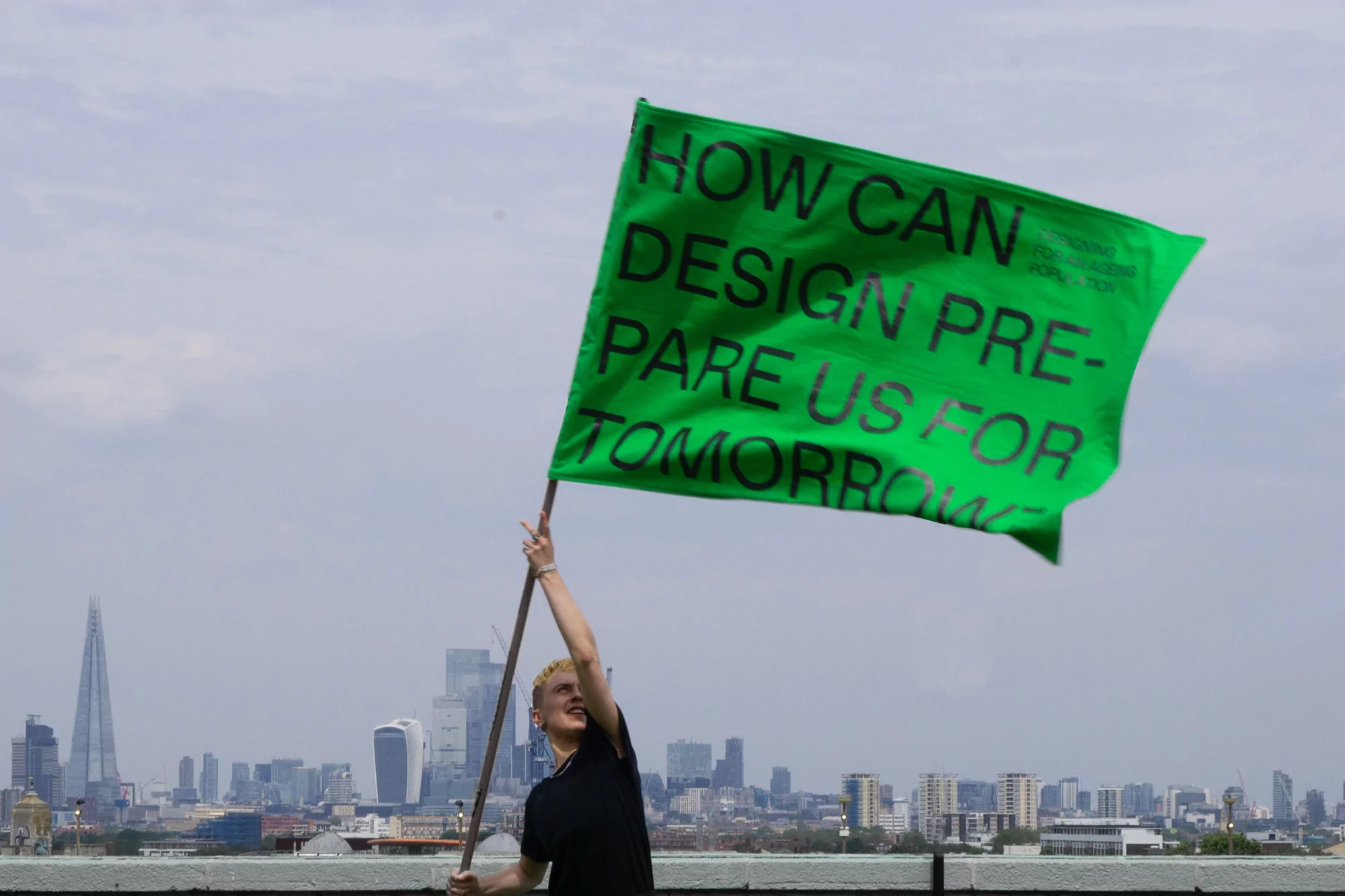 A young person holds a large green flag with black text that reads, 'How can design prepare us for tomorrow?' with a city skyline.
