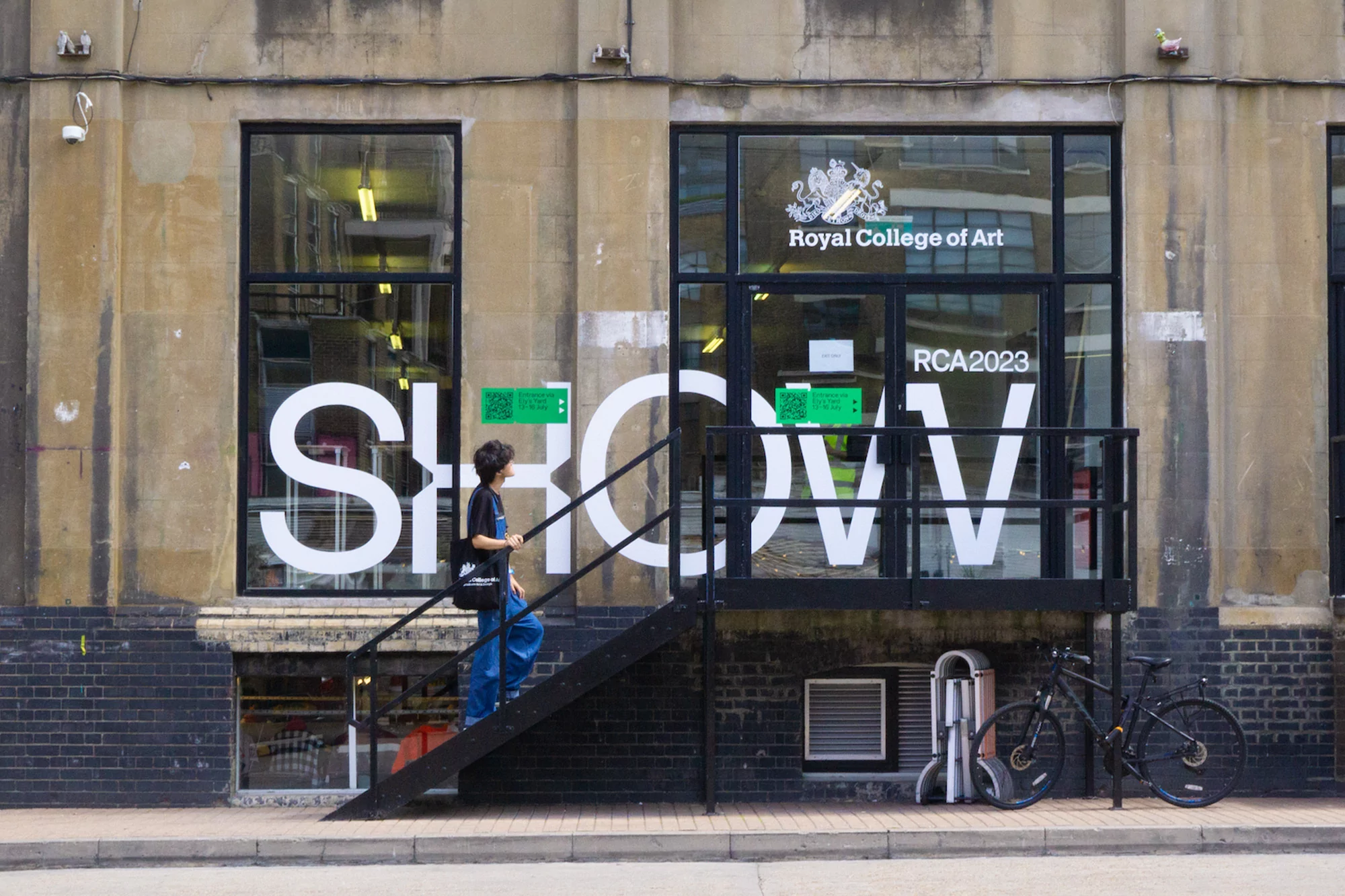 A woman walks up black stairs into the Royal College of Art SHOW 2023, beside a window with white SHOW letters on a beige stone building.
