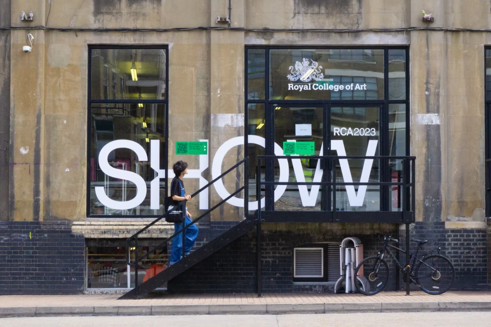 A woman walks up black stairs into the Royal College of Art SHOW 2023, beside a window with white SHOW letters on a beige stone building.