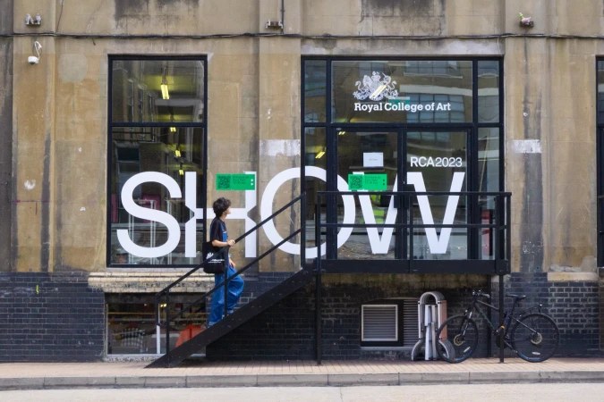 A woman walks up black stairs into the Royal College of Art SHOW 2023, beside a window with white SHOW letters on a beige stone building.