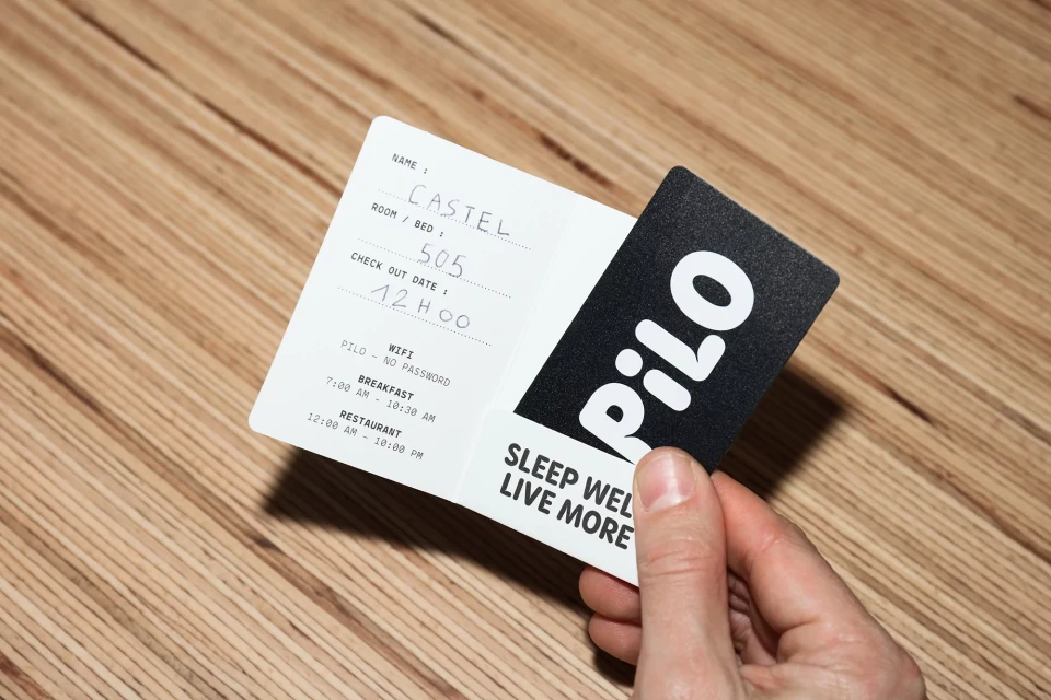 Hand holds a black and white hotel key card and a white information card on a wooden surface. Text includes hotel name, room number.