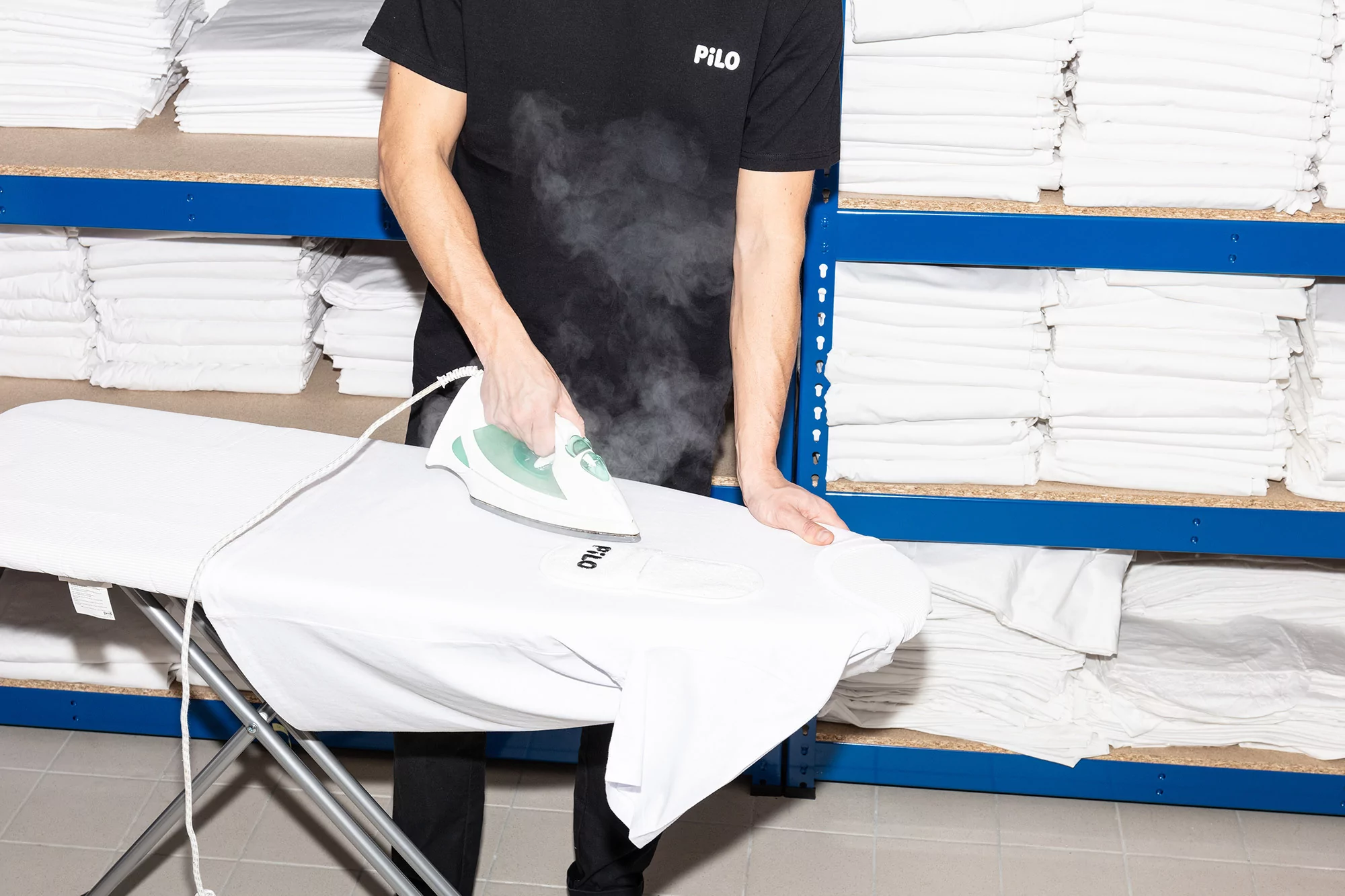 Adult male ironing a white shirt with 'PILO' logo on an ironing board with steam, with shelves of folded white linens in the background.