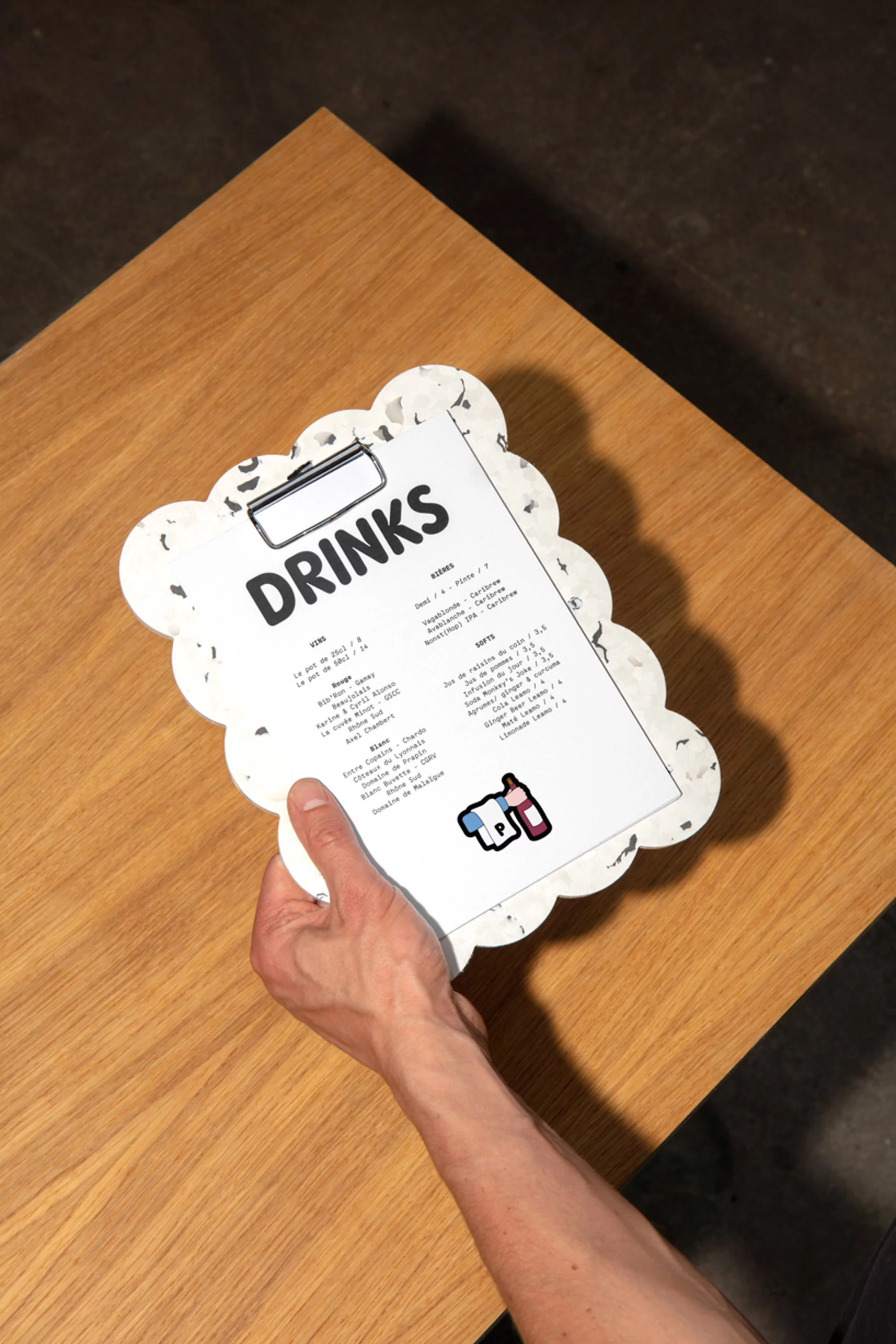 Person holding a drinks menu on a patterned clipboard at a bar or restaurant, with a table and dark background.