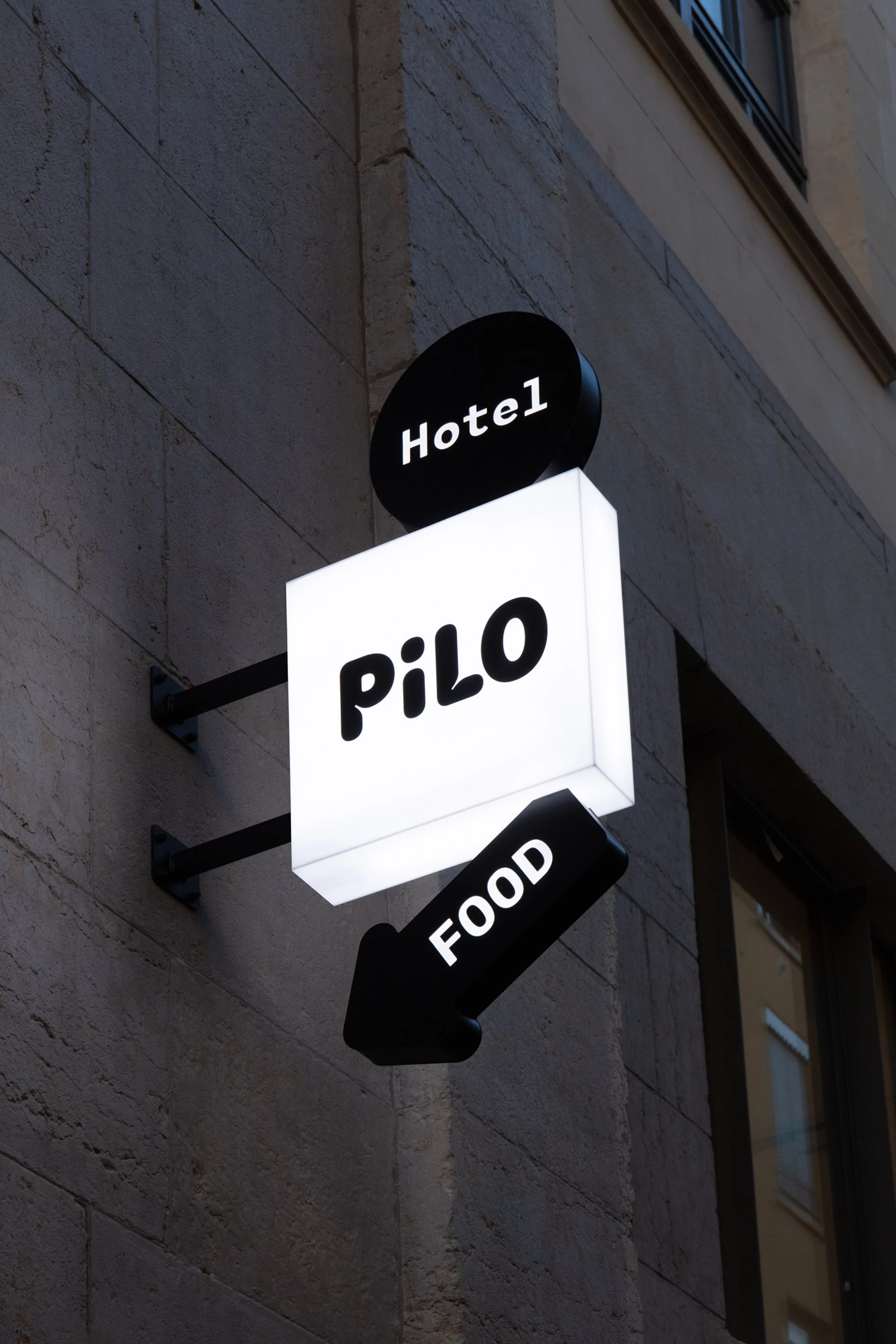 Illuminated sign on a building with the words 'Hotel,' 'PiLO,' and 'FOOD' in bold, rounded fonts with directional arrow.