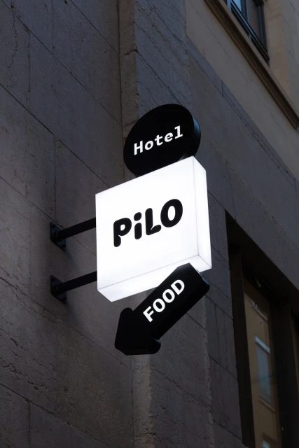 Illuminated sign on a building with the words 'Hotel,' 'PiLO,' and 'FOOD' in bold, rounded fonts with directional arrow.