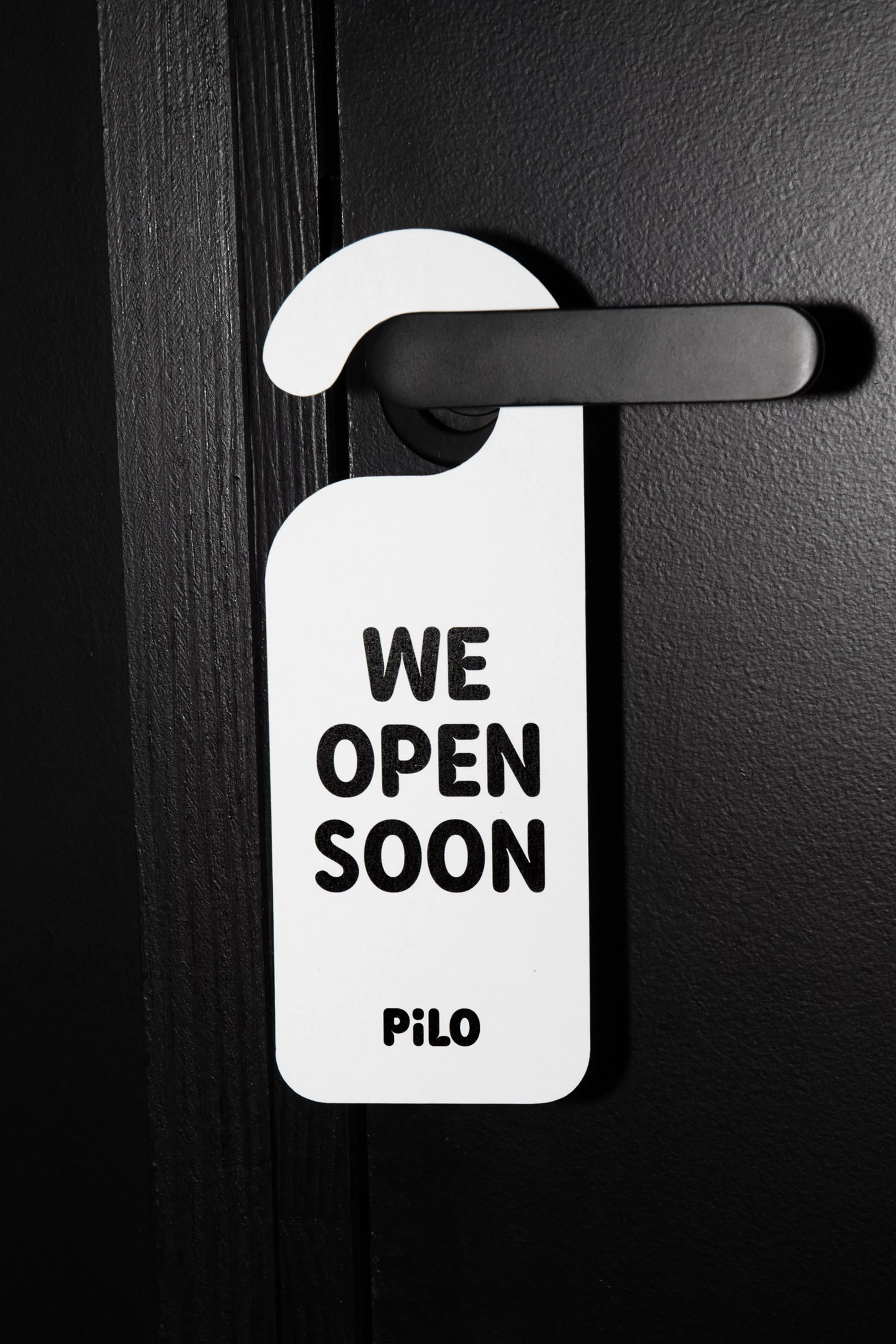Door hanger on a black door handle reads, "We open soon, PiLo." Advertisement for a hotel, business, or event opening.