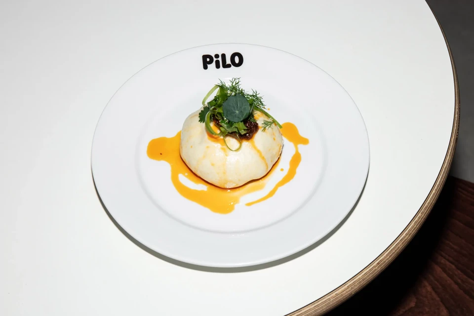 Overhead shot of a white plate with a savory dumpling topped with greens, drizzled with orange sauce on a white table. Food photography.