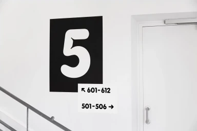 Monochrome architectural signage with a large white number five on a black rectangle next to directional wayfinding labels.