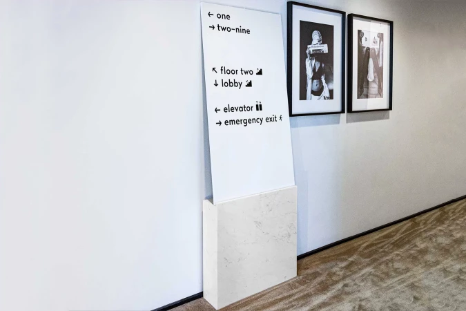 Interior shot of a marble-based sign with wayfinding text, accompanied by two framed black-and-white artworks hanging on a white wall.