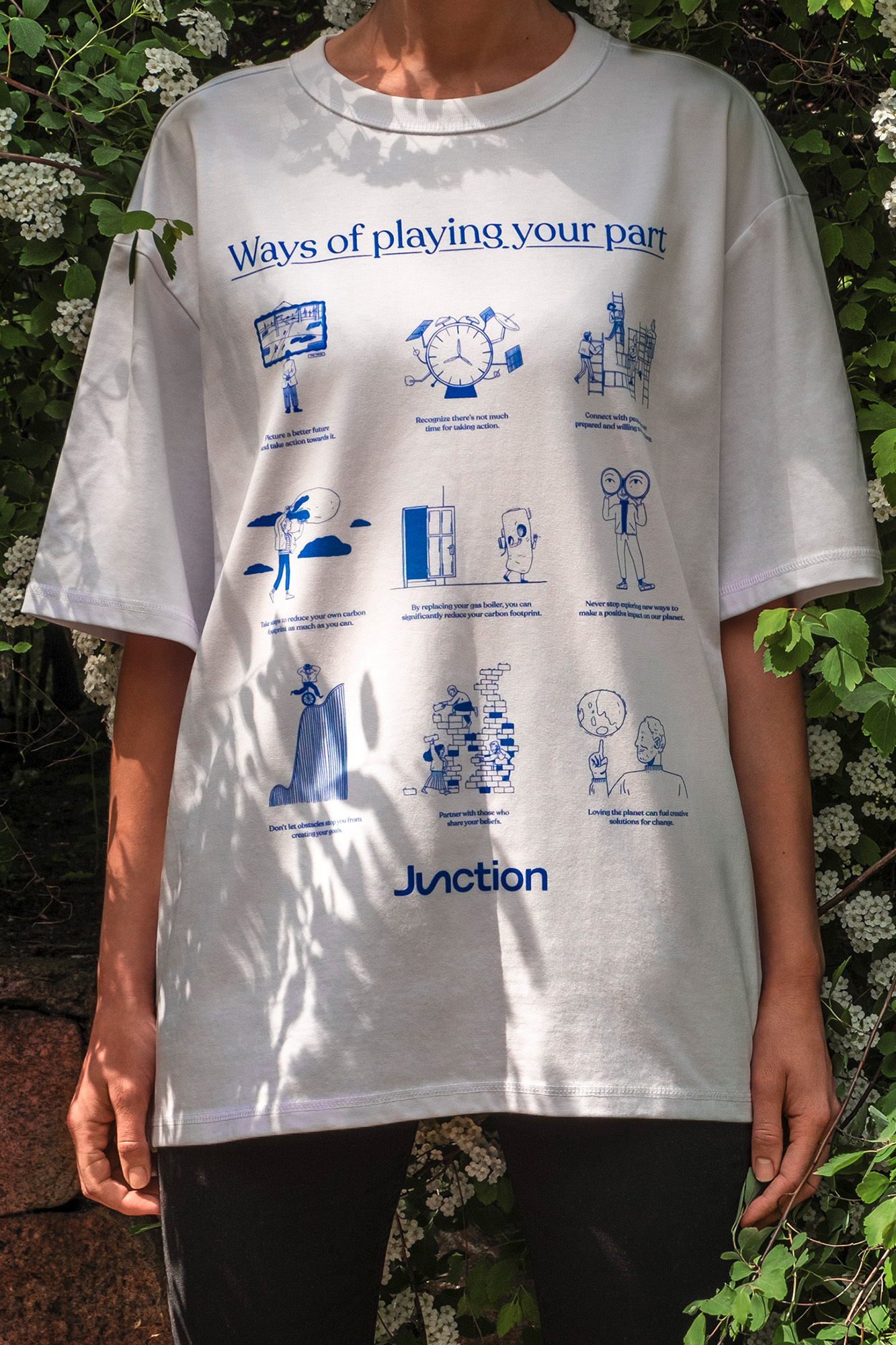 Person wearing a white t-shirt with blue illustrations of figures, objects, and text, standing outdoors surrounded by white flowers.