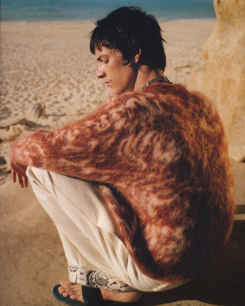 Young man in rust-colored sweater and white pants squats on sandy beach with ocean and sky backdrop in fashion style outdoor shot.