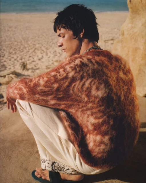 Young man in rust-colored sweater and white pants squats on sandy beach with ocean and sky backdrop in fashion style outdoor shot.