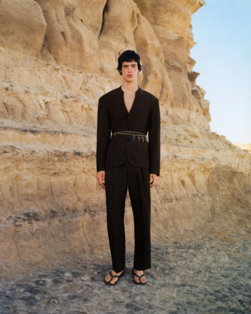 Man in dark suit and sandals poses against a light brown rock formation in a desert landscape, fashion editorial photography.