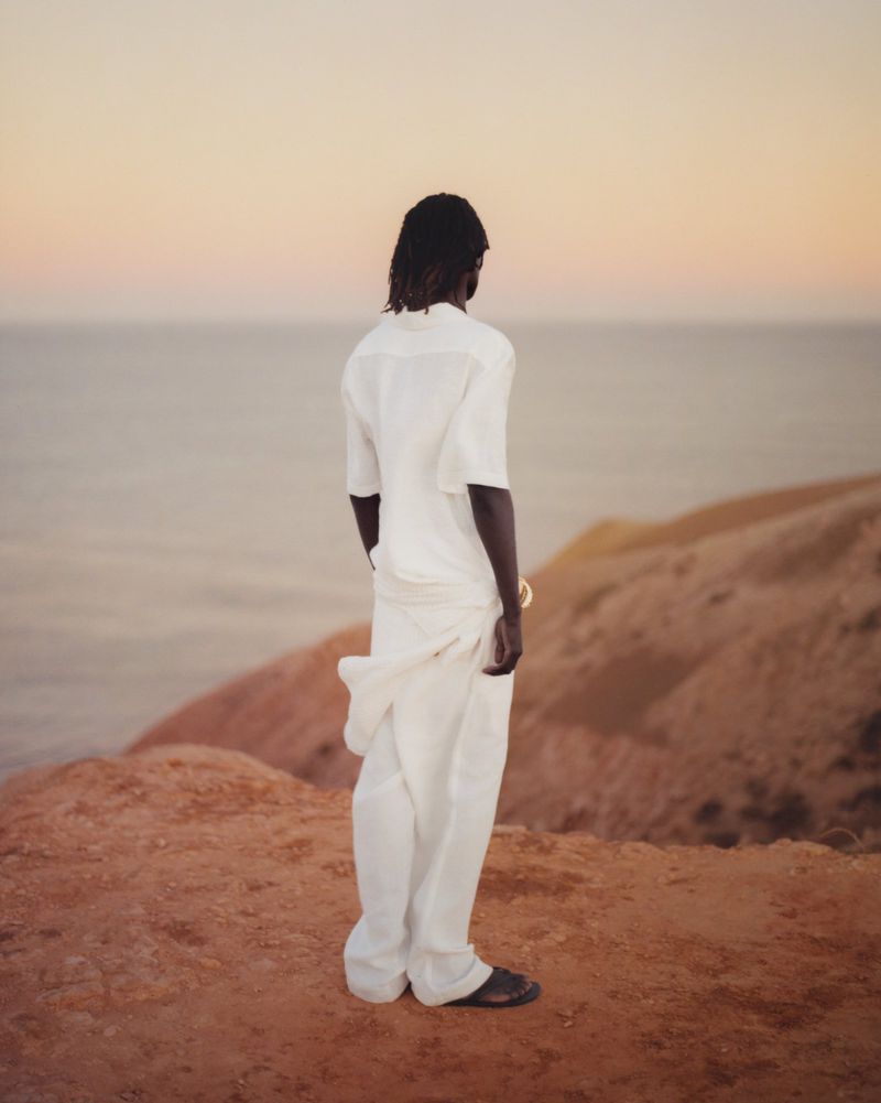 Man in a white linen outfit stands on a hillside overlooking the ocean during golden hour for a fashion or travel editorial.