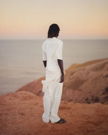 Man in a white linen outfit stands on a hillside overlooking the ocean during golden hour for a fashion or travel editorial.