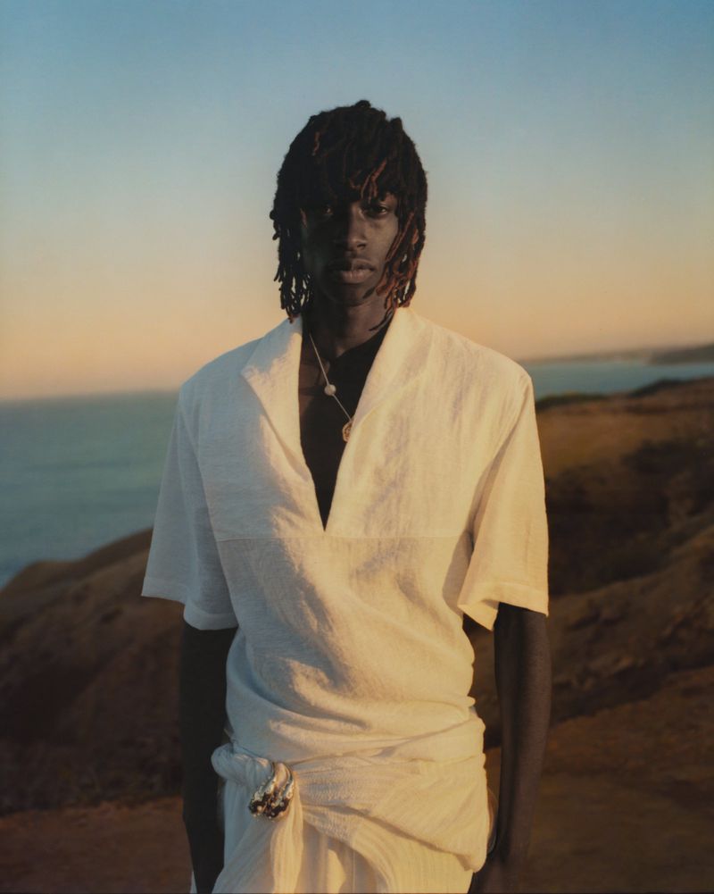 Portrait of an adult male in a white tunic standing on a hillside with a calm sea and a horizon visible under a blue and orange gradient sky.