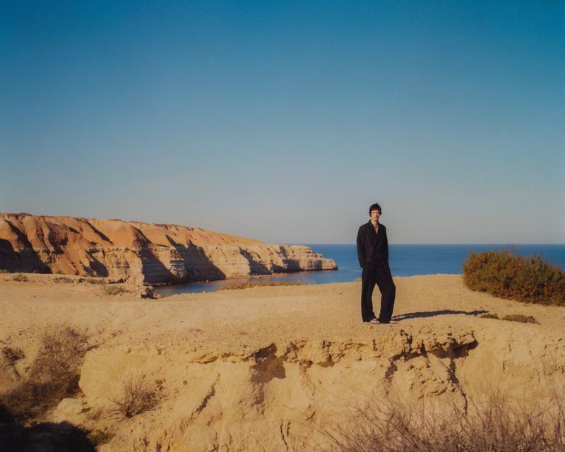 A full-length shot of a man standing on a cliff overlooking the ocean under a clear blue sky. He wears a dark outfit.