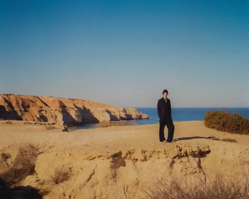 A full-length shot of a man standing on a cliff overlooking the ocean under a clear blue sky. He wears a dark outfit.