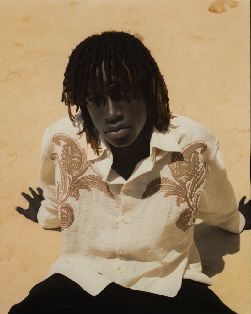 Portrait of a young adult with dreadlocks in a patterned shirt, posing on a sandy surface in natural light, captured in a close-up shot.