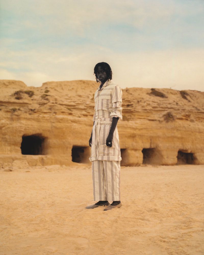 A person stands in a striped outfit in a desert landscape with rock formations and caves, under a blue sky with a neutral color grade.