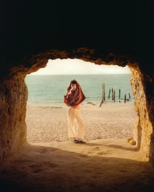 Woman standing on a beach, framed by a rock opening, wearing white pants and a rust-colored wrap with the ocean in the background.