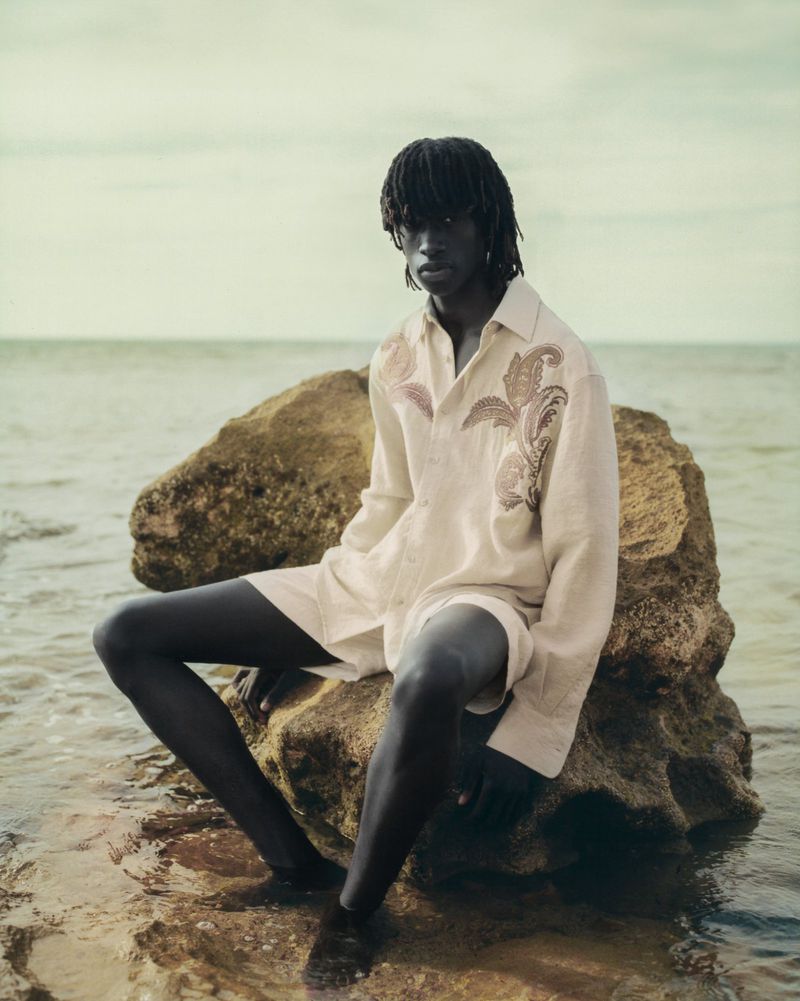Dark-skinned male model with dreadlocks sits on a large rock in water, wearing a patterned linen shirt and shorts against a cloudy sky.