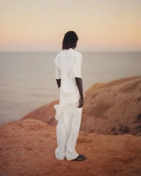 Man wearing a white shirt and trousers standing on a coastal cliff edge at sunset for COMMAS Pre Fall 2025 editorial