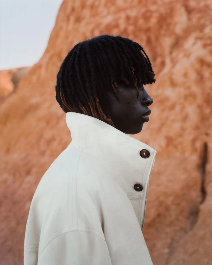 Profile portrait of a Black man with dreadlocks wearing a cream high-collar jacket against a textured red-brown earth background