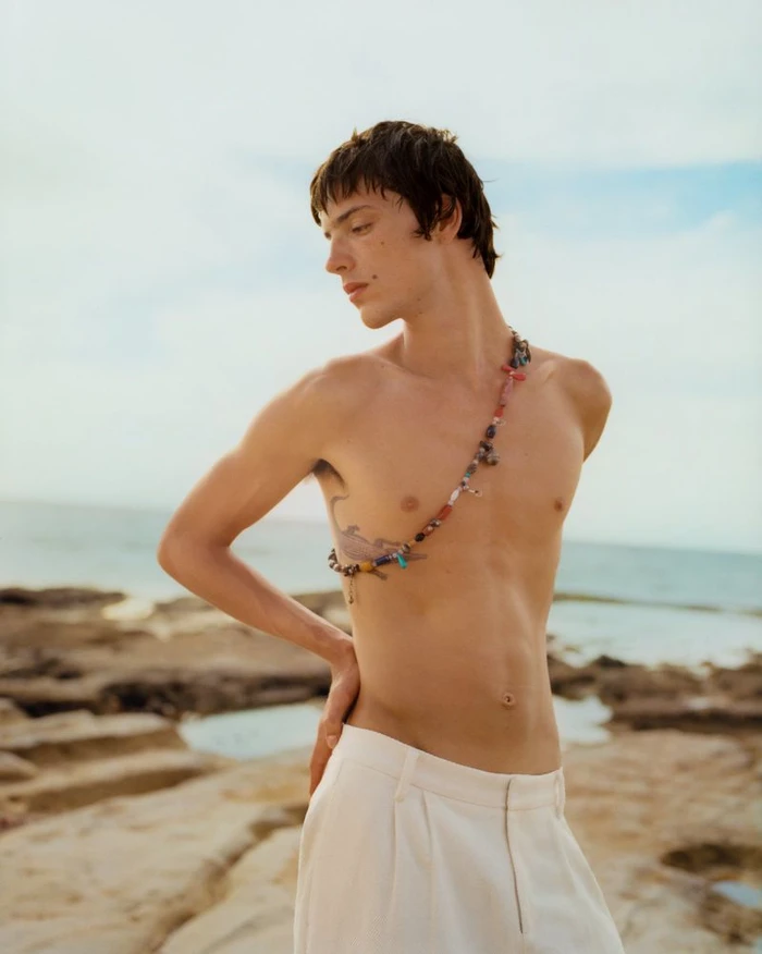 Shirtless male model wearing white trousers and a beaded body chain standing by a rocky coast.