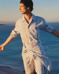 Model wearing a blue and white striped shirt and shorts against an ocean background for COMMAS Pre Fall 2025.