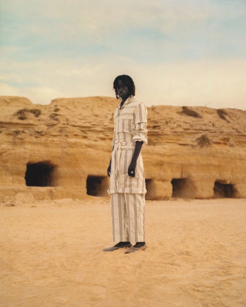 A male model standing in a sandy desert environment wearing a striped matching shirt and trousers ensemble.