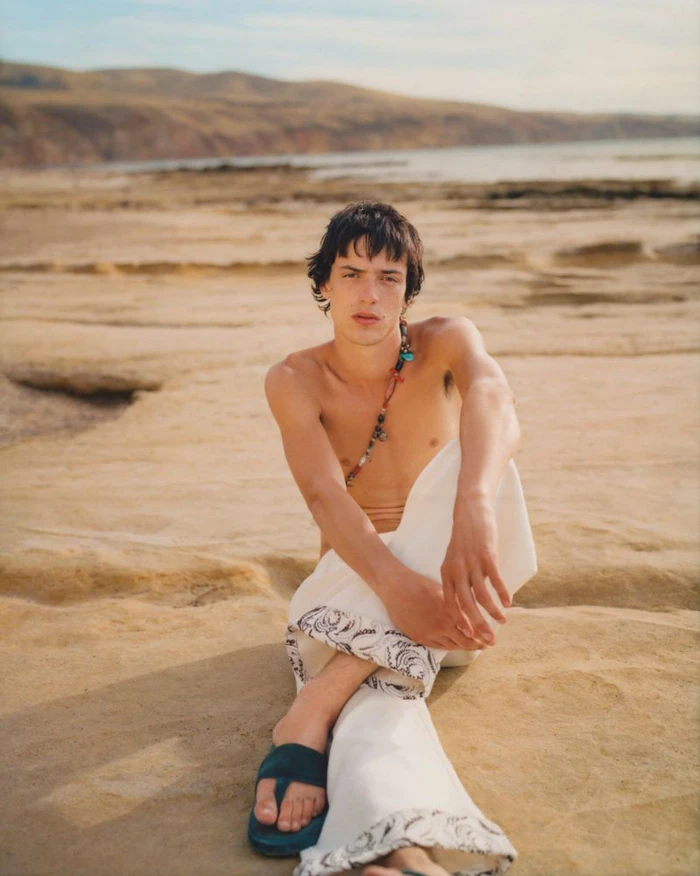Shirtless male model sitting on sandy rocky coastline wearing patterned trousers and beaded necklace