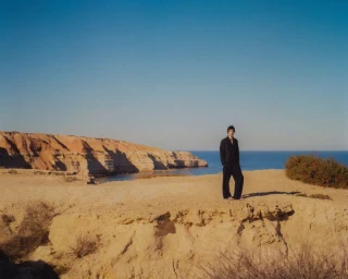 A man standing on a sandy coastal cliff overlooking the ocean under a clear blue sky, photographed for COMMAS Pre Fall 2025.