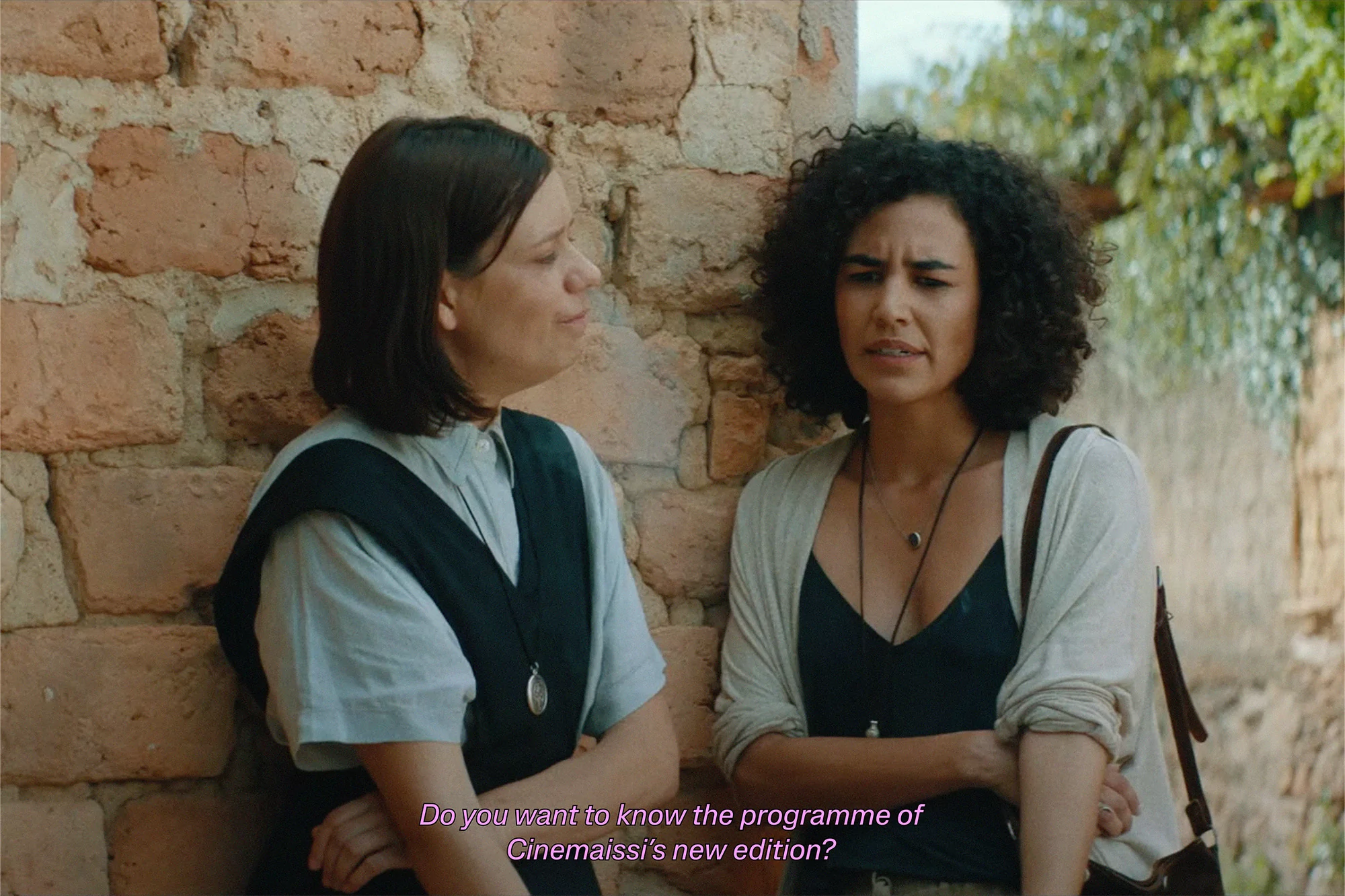 Two women in casual attire stand near a textured brick wall. One wears a religious-style outfit with a pendant, while the other has curly hair.